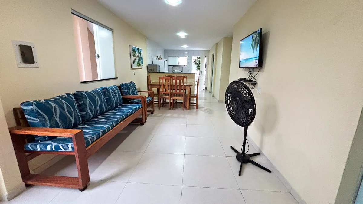 Sala integrada ampla com sofa de madeira, mesa de jantar, TV e cozinha ao fundo
