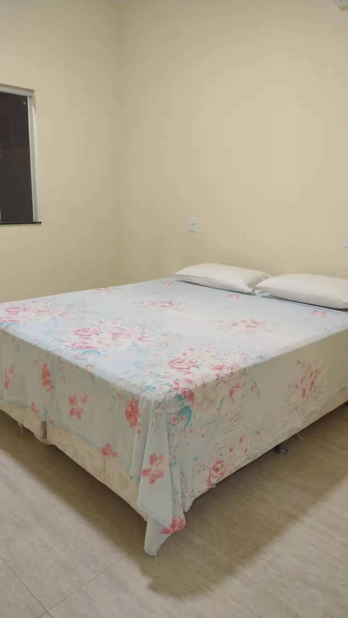Quarto com cama de casal, roupa de cama floral e janela lateral.