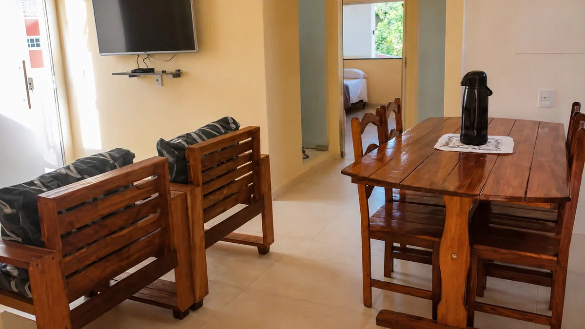 Sala de estar e jantar com sofa de madeira, mesa, TV e acesso aos quartos.