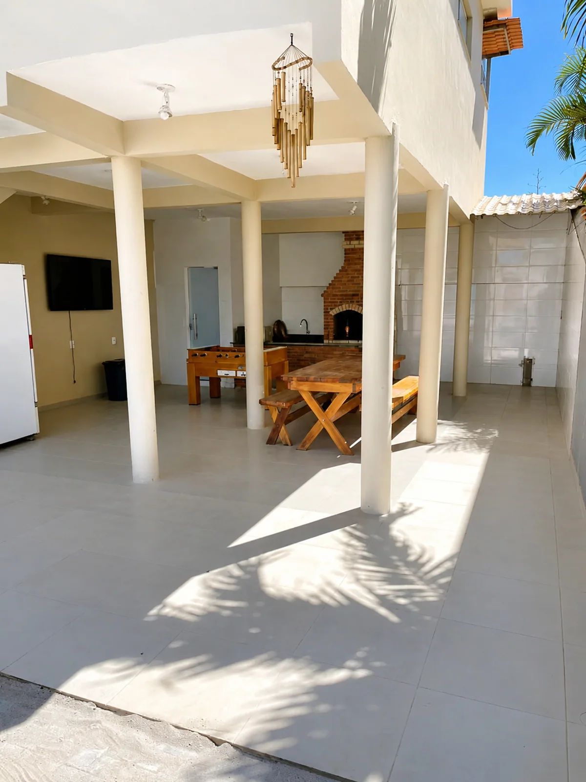 Area externa coberta com mesa de madeira, churrasqueira de tijolos e mesa de jogos.