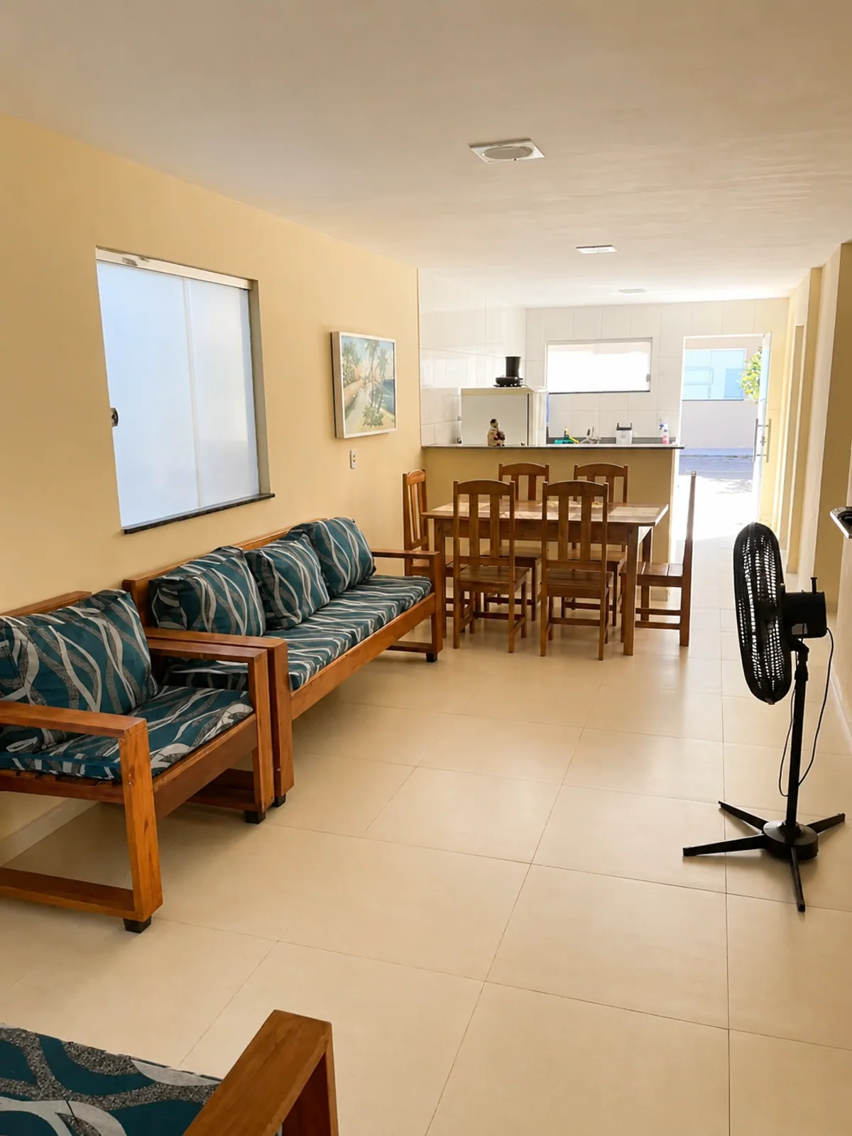 Sala de estar e jantar com sofa, mesa de madeira, ventilador e cozinha ao fundo.